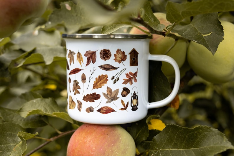 May include: A white enamel mug with a silver rim and a handle, featuring a repeating pattern of brown and orange autumn leaves, acorns, a small wooden birdhouse, and a vintage-style lantern printed on the exterior surface of the cup.