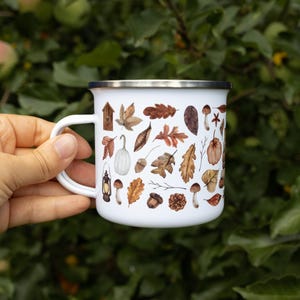 May include: A white enamel mug with a silver rim featuring a repeating pattern of brown autumn leaves, acorns, pinecones, mushrooms, a small birdhouse, and a vintage lantern. The mug has a curved handle and a smooth, glossy finish throughout.
