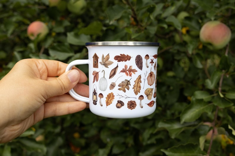 May include: A white enamel mug with a silver rim featuring a repeating pattern of brown autumn leaves, acorns, pinecones, mushrooms, a small birdhouse, and a vintage lantern. The mug has a curved handle and a smooth, glossy finish throughout.