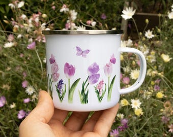 May include: A white enamel mug featuring a colorful botanical print of purple tulips, pink hyacinths, and small butterflies. The mug has a smooth white finish, a curved handle on the right side, and a thin silver-toned metal rim around the top edge.