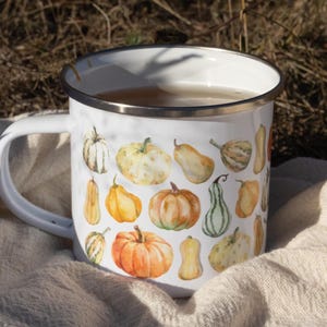 Puede incluir: Una taza de esmalte blanco con un colorido estampado de acuarela de varias calabazas en tonos naranja, amarillo y verde. La taza tiene un acabado blanco liso, un borde de metal plateado y un asa curva en el lado izquierdo.