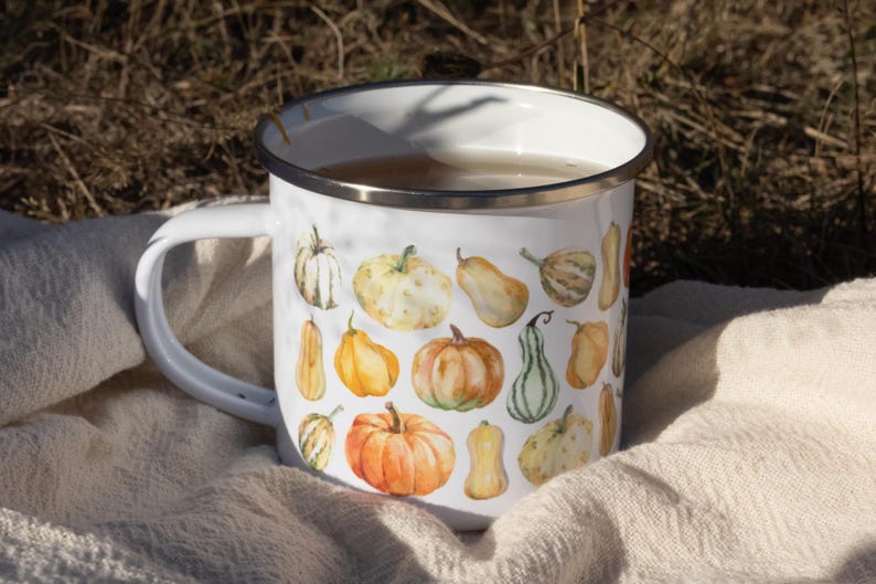 Puede incluir: Una taza de esmalte blanco con un colorido estampado de acuarela de varias calabazas en tonos naranja, amarillo y verde. La taza tiene un acabado blanco liso, un borde de metal plateado y un asa curva en el lado izquierdo.