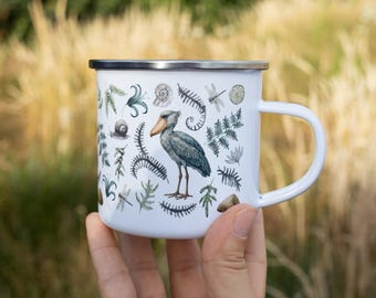 May include: A white enamel mug featuring a detailed illustration of a shoebill stork surrounded by various botanical elements, ferns, dragonflies, and a snail. The mug has a silver-colored metal rim and a matching white handle on the right side.