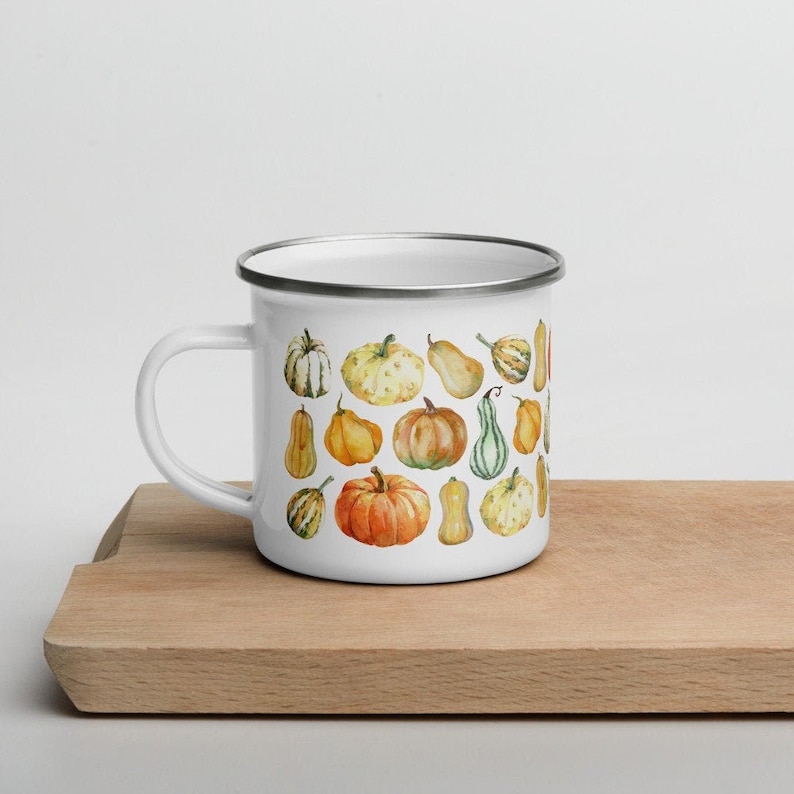 Puede incluir: Una taza de esmalte blanco con borde plateado que presenta un estampado de acuarela de varias calabazas en tonos naranja, amarillo, verde y crema. La taza tiene un asa curva en el lado izquierdo y un acabado suave y brillante.