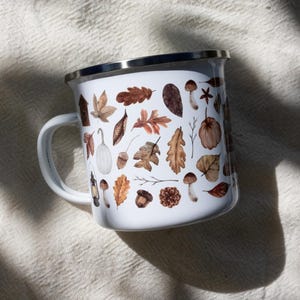 May include: A white enamel mug with a silver rim and a handle, featuring a repeating pattern of watercolor-style autumn elements including brown oak leaves, acorns, pinecones, mushrooms, a small white pumpkin, and a vintage-style lantern.