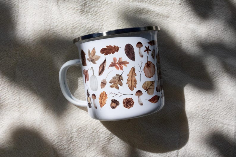 May include: A white enamel mug with a silver rim and a handle, featuring a repeating pattern of watercolor-style autumn elements including brown oak leaves, acorns, pinecones, mushrooms, a small white pumpkin, and a vintage-style lantern.