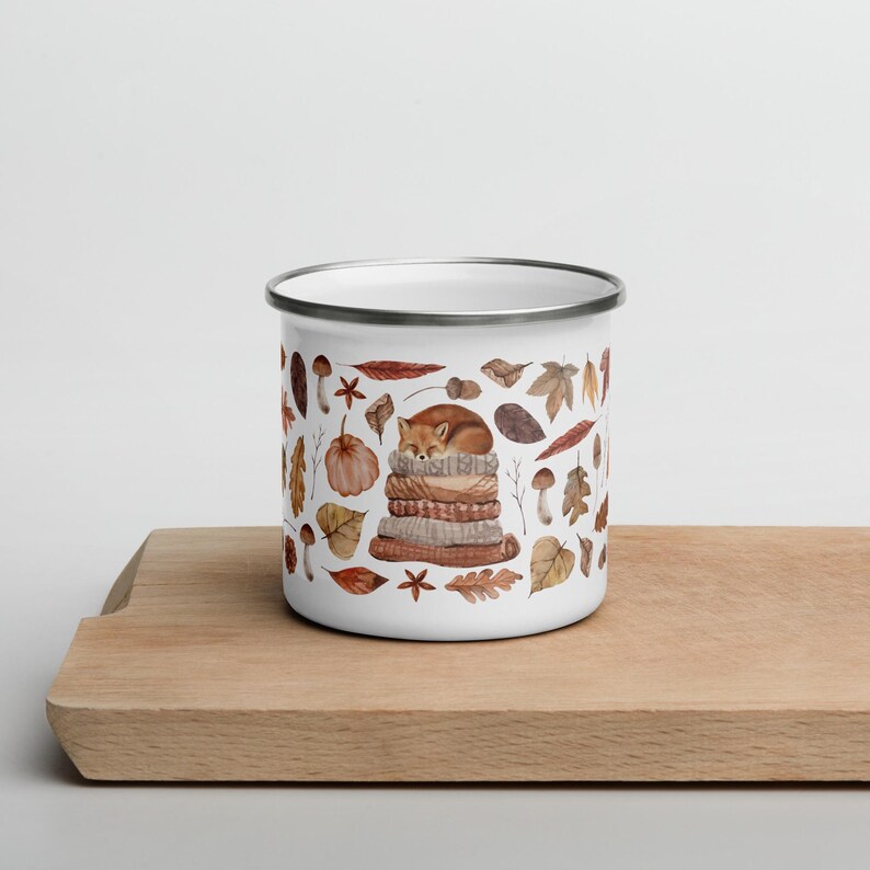 White enamel mug with a silver rim featuring a cute illustrated fox sleeping on a stack of cozy blankets surrounded by autumn leaves, pumpkins, mushrooms, and fall elements, displayed on a wooden board against a light background.