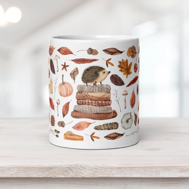 White ceramic mug featuring a cute illustrated hedgehog sitting on a stack of cozy blankets surrounded by autumn leaves, pumpkins, mushrooms, and fall elements, displayed on a wooden white board against a light background.