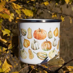 Puede incluir: Una taza de esmalte blanco con borde plateado, decorada con un patr&oacute;n repetitivo de estilo acuarela de varias calabazas y calabacines naranjas, amarillos y verdes de diferentes formas y tama&ntilde;os, sobre un fondo blanco.