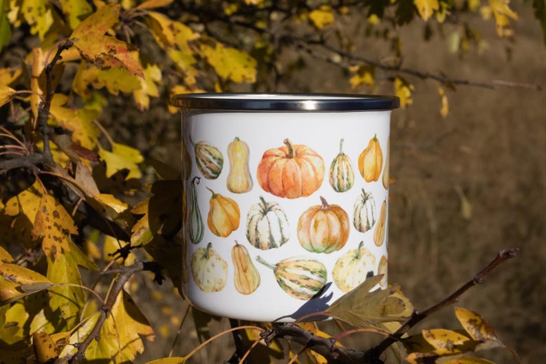 Puede incluir: Una taza de esmalte blanco con borde plateado, decorada con un patr&oacute;n repetitivo de estilo acuarela de varias calabazas y calabacines naranjas, amarillos y verdes de diferentes formas y tama&ntilde;os, sobre un fondo blanco.