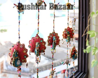 May include: A colorful string of peacock-shaped fabric ornaments hanging in a window. The ornaments are made of beige, red, and green fabric and are decorated with sequins and beads. The string is hanging in front of a window with a view of a beach and ocean.