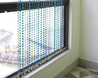 May include: Turquoise beaded curtain hanging in front of a window. The curtain is made of strands of turquoise beads, with a metallic top and bottom. The window has a black frame and is partially open, with a view of the outside.