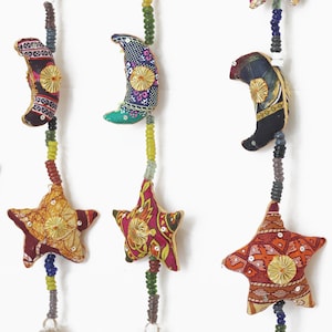 Peut inclure: Une guirlande colorée de tissu en forme d'étoiles et de croissants de lune avec des cloches au bas. Les étoiles et les lunes sont faites de tissu patchwork avec des accents dorés. La guirlande est faite de perles de différentes couleurs.