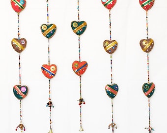 May include: Five strands of colorful heart-shaped fabric ornaments hang from a white background. Each strand is adorned with beads and a bell at the bottom. The hearts are decorated with sequins, embroidery, and gold trim.