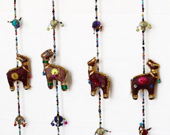 May include: A set of six colorful fabric animal ornaments hanging from strings of beads. Each ornament is a camel with gold trim and a bell at the bottom of each string.