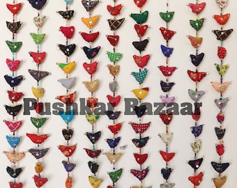 May include: Seven colorful fabric bird garlands with bells at the bottom. Each garland has 15 small fabric birds in various colors and patterns. The garlands are hanging from hooks on a white wall. The text "Pushkar Bazaar" is visible in the center of the image.