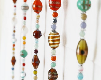 May include: Five strands of colorful beads hang from a white background. The beads are made of glass and stone and come in a variety of shapes and sizes. The beads are strung on a clear thread.