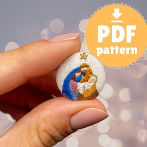 Puede incluir: Un botón de tela blanco con una imagen bordada de la Sagrada Familia. La imagen presenta a María, José y el niño Jesús en una gama de colores azul, rosa y dorado. Una estrella dorada está bordada sobre la familia.