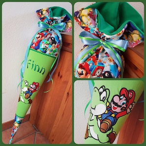 Könnte beinhalten: Eine grüne, kegelförmige Schultüte mit einem farbenfrohen Mario Bros.-Aufdruck. Die Tüte zeigt ein gesticktes Bild von Mario und Yoshi mit dem Namen "Finn" in Grün. Sie ist mit blauen und grünen Bändern gebunden.