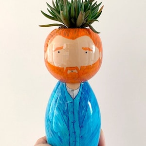 May include: A hand-painted ceramic planter shaped like a person with a green succulent plant growing from the top of the head. The planter is painted with a light blue jacket, a white shirt, and a red beard. The head of the planter is painted with a light orange skin tone and brown eyes.