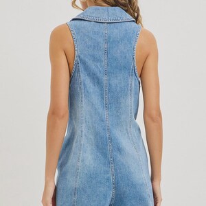 May include: Light blue denim romper with a collared neckline and sleeveless design. The romper features a button-up front and a tailored fit, showcasing a classic denim aesthetic. The shorts end at mid-thigh.