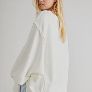 May include: Cream-colored oversized sweatshirt with a relaxed fit. Features a wide neckline, dropped shoulders, and visible stitching details. The long sleeves and ribbed hem add to the casual style. The model is wearing denim shorts.