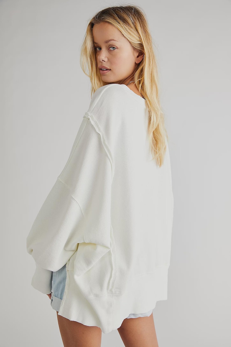 May include: Cream-colored oversized sweatshirt with a relaxed fit. Features a wide neckline, dropped shoulders, and visible stitching details. The long sleeves and ribbed hem add to the casual style. The model is wearing denim shorts.