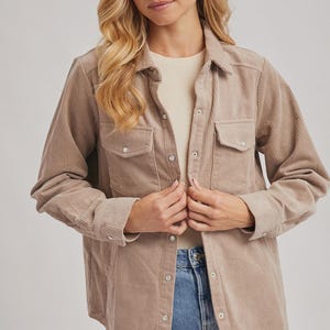 May include: A tan corduroy shirt with two chest pockets and silver snap closures. The shirt is worn over a cream-colored top and paired with denim shorts. The corduroy fabric has vertical ridges.