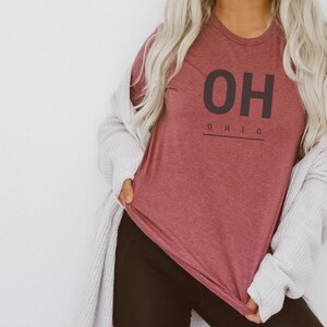 May include: A maroon t-shirt with the text "OH" in black, followed by "OHIO" in smaller black text underneath. The shirt is being worn by a person with long blonde hair.