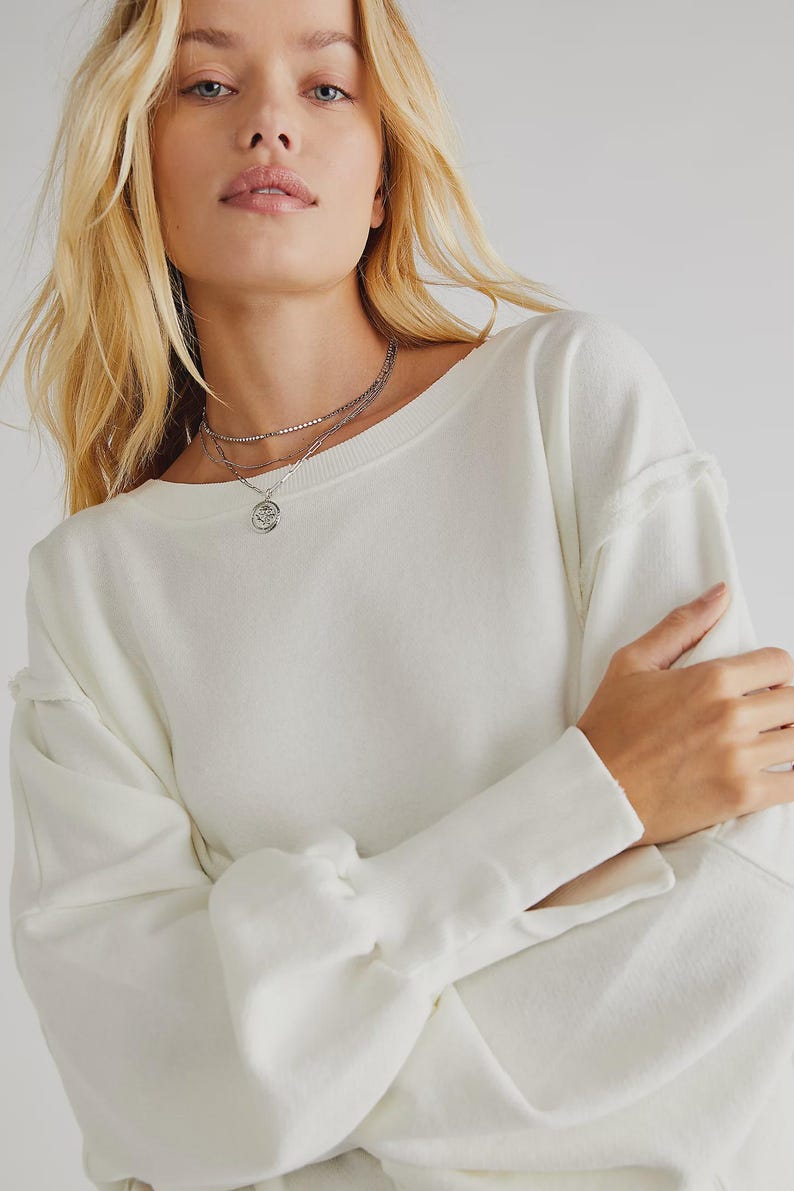 May include: Cream-colored long-sleeve sweatshirt with a wide neckline and raw edge detailing. The model is wearing a layered silver necklace with a pendant. The sleeves have a gathered design at the wrist.