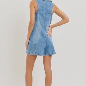 May include: Light blue denim romper with a collared neckline and sleeveless design. The romper features a button-down front and a tailored fit. The shorts have a slight flare. The model is wearing beige sandals.