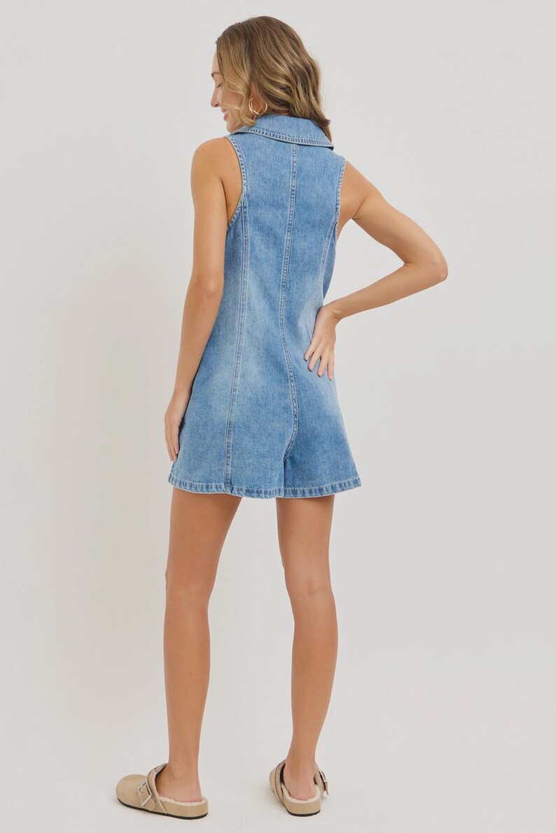 May include: Light blue denim romper with a collared neckline and sleeveless design. The romper features a button-down front and a tailored fit. The shorts have a slight flare. The model is wearing beige sandals.