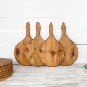 May include: Four wooden pizza paddles with handles. The paddles are light brown and have a natural wood finish. They are arranged in a row on a white surface.