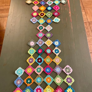 May include: A colorful, handmade crochet table runner. The runner is made of numerous square motifs, each featuring a flower design in various colors, including pink, yellow, blue, and green. The runner is laid out on a dark green surface.