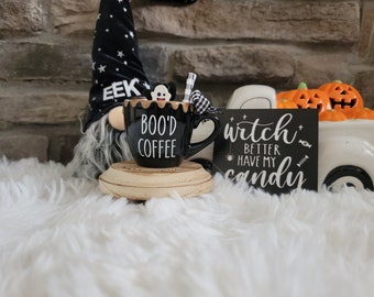 May include: A Halloween-themed display featuring a black coffee mug with "BOO'D COFFEE" text, a gnome, and a sign that reads "witch better have my candy". The mug sits on a wooden coaster, and the scene includes decorative pumpkins.