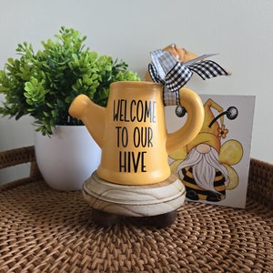 May include: Yellow ceramic watering can with a black and white checkered bow and the text "Welcome to our Hive". The watering can is sitting on a wooden base and a small bee gnome card is in the background.