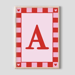 May include: A framed art print with a large red letter "A" on a pale pink background. The border has a red and pink chequered pattern, with small red hearts in each corner. The frame is a natural wood colour.