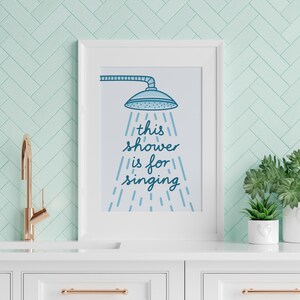 May include: A framed print with the text "this shower is for singing" in blue script, featuring a showerhead illustration. The print is displayed on a white cabinet with a gold tap and potted plants, against a light green tiled wall.