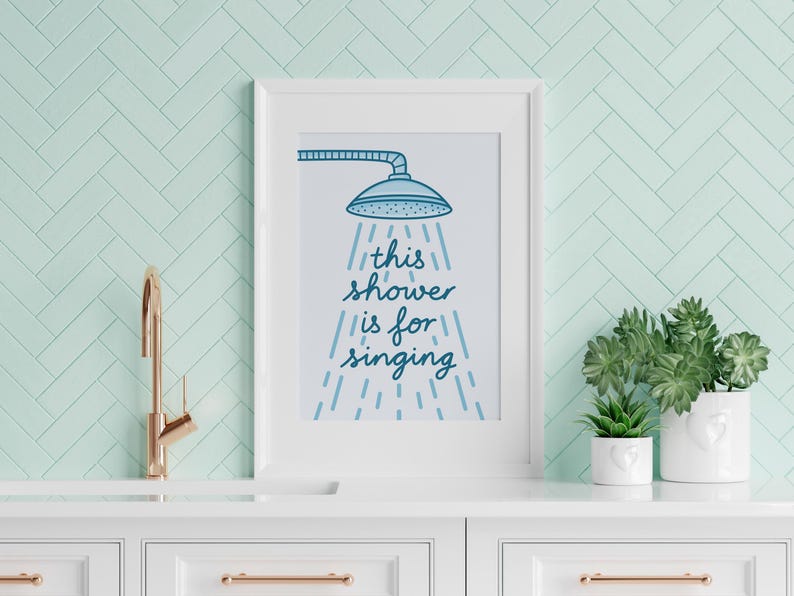 May include: A framed print with the text "this shower is for singing" in blue script, featuring a showerhead illustration. The print is displayed on a white cabinet with a gold tap and potted plants, against a light green tiled wall.