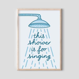 May include: A framed art print with a blue showerhead illustration and the text "this shower is for singing". The artwork uses a light blue and white colour scheme. Ideal for bathroom decor, the print is in a wooden frame.