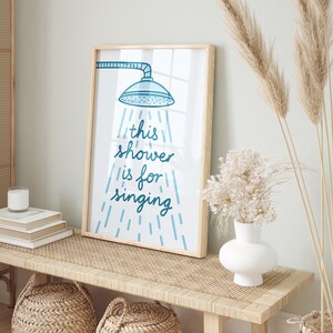 May include: A framed print with the text "this shower is for singing" in blue script. The artwork features a blue showerhead illustration with water droplets. The frame is light wood, and the print is displayed on a woven bench.