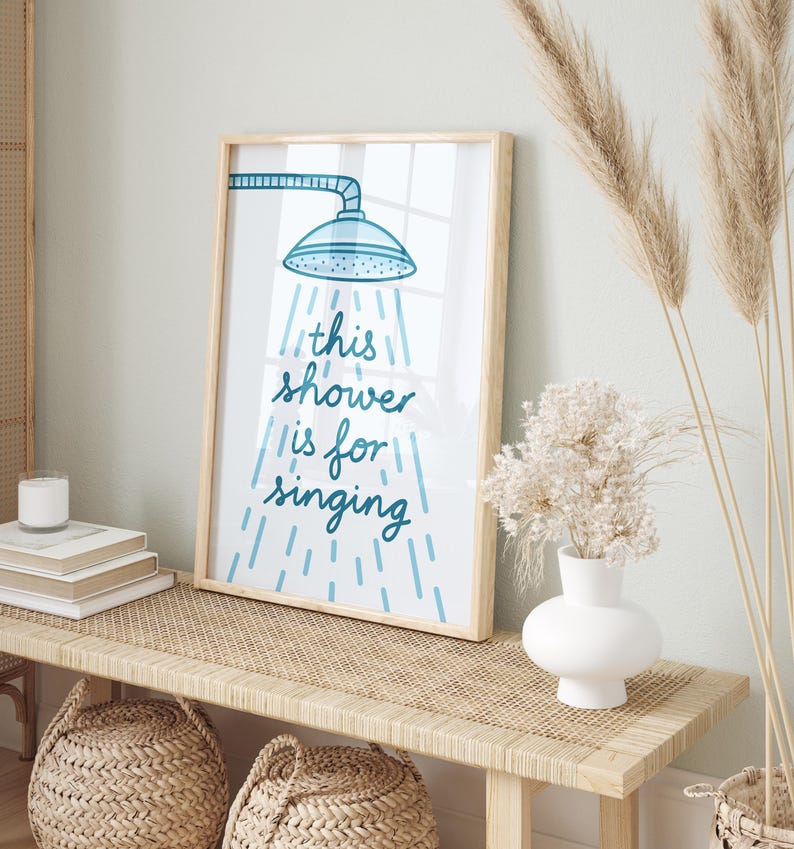 May include: A framed print with the text "this shower is for singing" in blue script. The artwork features a blue showerhead illustration with water droplets. The frame is light wood, and the print is displayed on a woven bench.