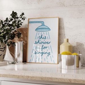 May include: A framed print with a gold border shows a blue showerhead illustration and the text "this shower is for singing" in blue. The print is on a white countertop with other decorative items.