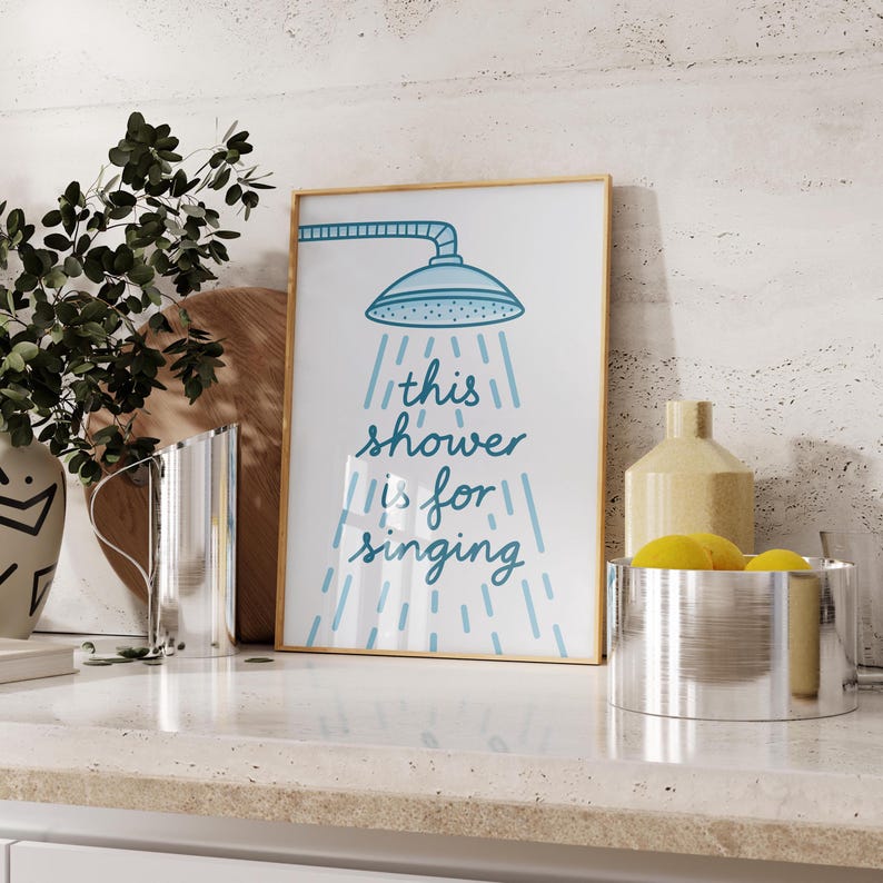 May include: A framed print with a gold border shows a blue showerhead illustration and the text "this shower is for singing" in blue. The print is on a white countertop with other decorative items.