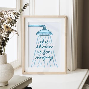 May include: A framed print with a light wood frame. The print features a blue showerhead illustration with the words "this shower is for singing" in a handwritten style. The artwork is displayed on a white surface.