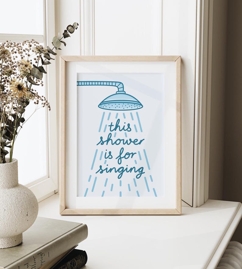 May include: A framed print with a light wood frame. The print features a blue showerhead illustration with the words "this shower is for singing" in a handwritten style. The artwork is displayed on a white surface.