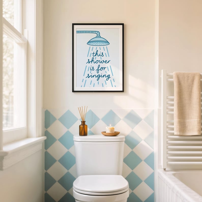 May include: A bathroom interior with a framed print displaying a showerhead and the words "this shower is for singing." The room includes blue and white diamond tiles, a white toilet, and a window. A towel is on a radiator.