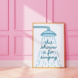 May include: A framed art print with a gold-coloured frame. The print features a blue showerhead illustration with the text "this shower is for singing" in a matching blue font. The background is white.