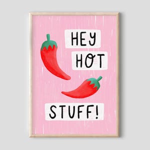 May include: A framed print with a pink background, featuring two red chillies with green stems. The words "HEY HOT STUFF!" are written in black on white rectangular blocks. The artwork has a playful, illustrative style.