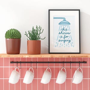 May include: A black-framed print with the text "this shower is for singing" is displayed above a shelf with two potted plants and five white mugs. The print features a blue showerhead illustration. The shelf is above a pink tiled wall.
